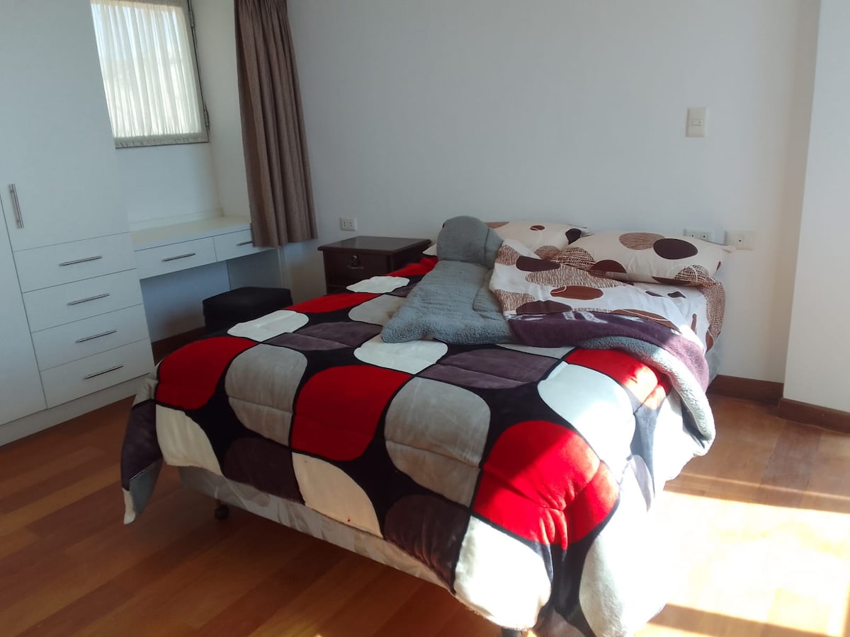 A bright private room features a spacious bed covered with a patterned blanket. A large window allows natural light, illuminating the wooden floor. A desk and chair are positioned against one wall, alongside a dresser providing storage space.