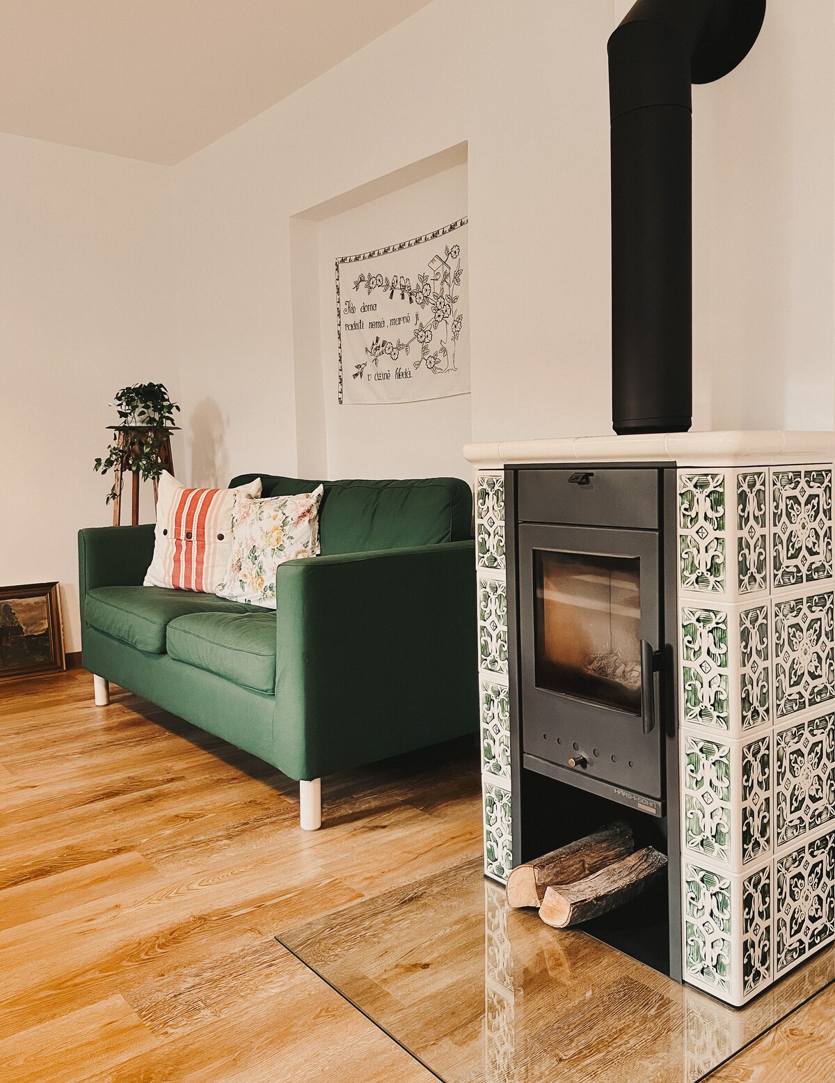 A cozy seating area features a green couch adorned with decorative cushions. Nearby, a tiled wood stove provides warmth and ambiance, with logs neatly placed for convenience. Light hardwood flooring complements the bright and inviting atmosphere.