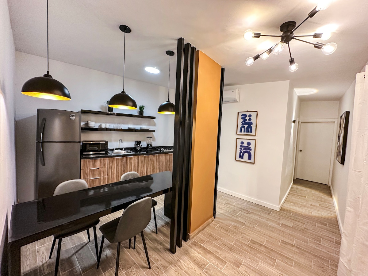A modern kitchen area features a sleek black dining table surrounded by four gray chairs. Overhead, pendant lights illuminate a well-equipped kitchen with contemporary appliances, including a refrigerator and microwave. A stylish partition separates the dining area from the hallway.