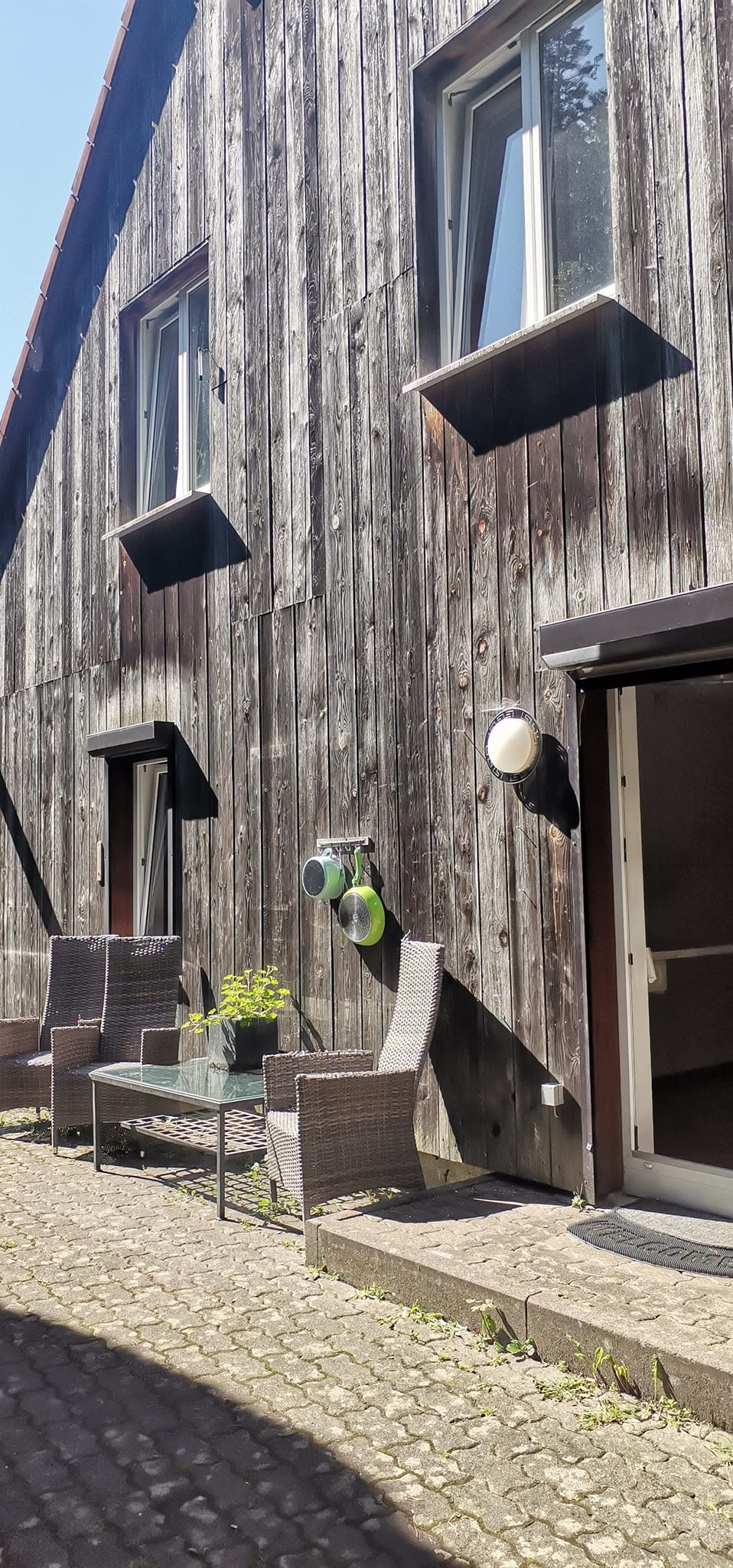 An exterior view of the building features rustic wooden cladding with several windows. Two wicker chairs and a small table are placed on cobblestone flooring, creating a cozy outdoor seating area. A green planter adds a touch of nature to the space.