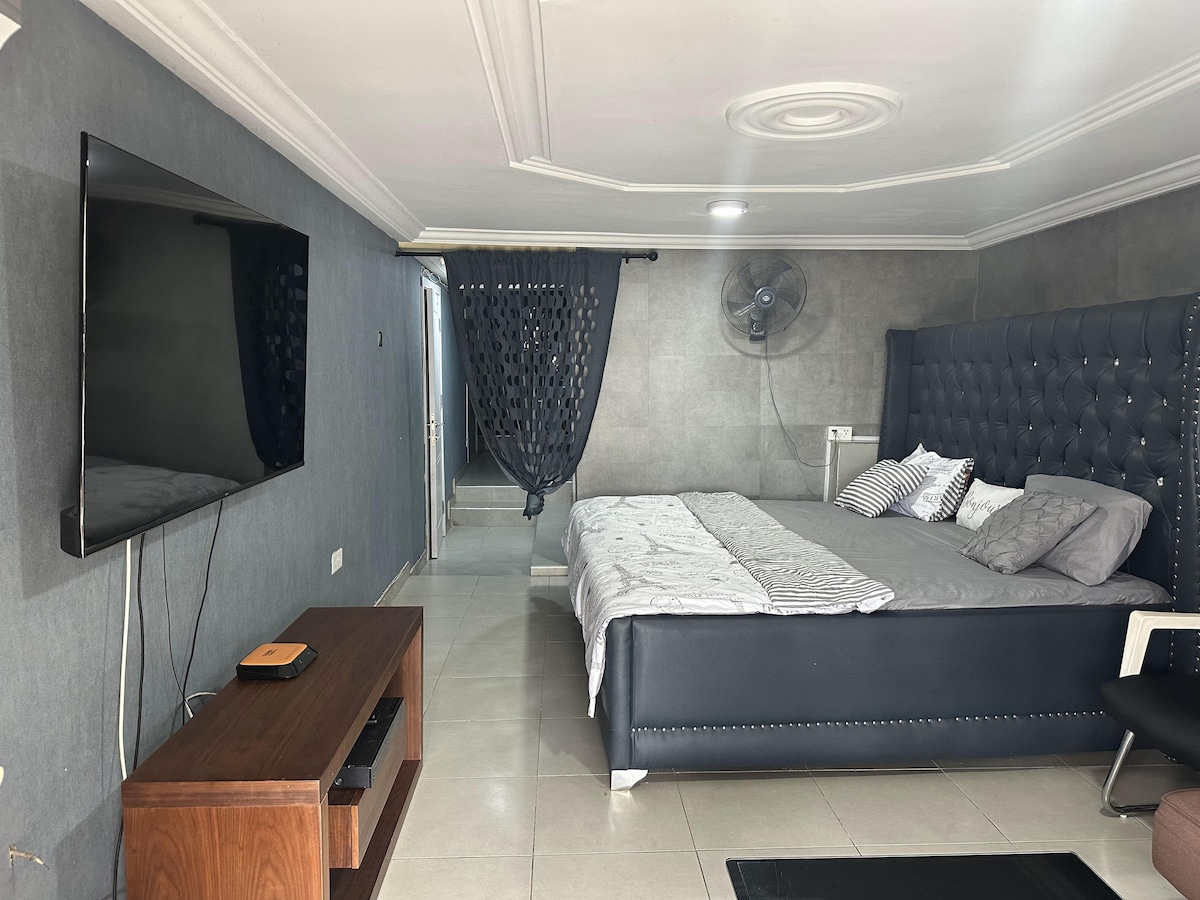 A modern sleeping area features a spacious bed with decorative pillows. A large wall-mounted television is positioned across from the bed, complemented by a wooden entertainment unit. Gray walls and polished tiles contribute to the contemporary aesthetic. A fan is mounted above for added comfort.