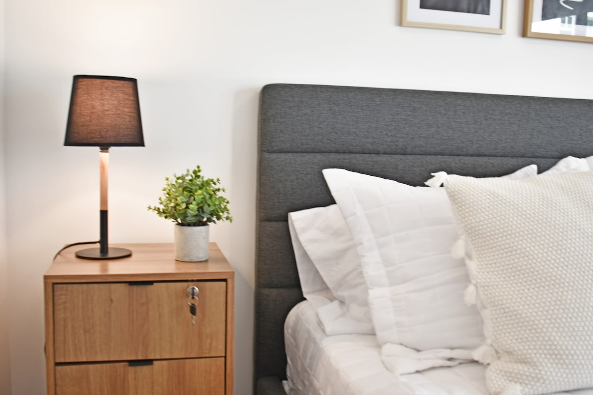 A serene bedroom corner features a gray upholstered headboard and a neatly made spacious bed with white bedding. A bedside table holds a small potted plant and a stylish lamp, offering a peaceful atmosphere with soft lighting.