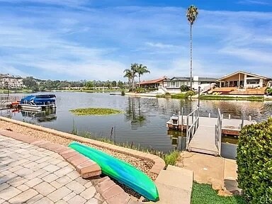 A serene lakefront view is captured, showcasing calm waters reflecting the sky. A partially visible dock extends from the shoreline, while a kayak rests on the paved area. Palm trees provide a touch of greenery against the backdrop of neighboring homes.