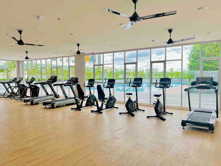 A well-equipped fitness center features various cardio machines, including treadmills and stationary bikes. Large windows provide ample natural light and offer views of the outdoor swimming pool. The wooden flooring adds warmth to the modern space, while ceiling fans contribute to air circulation.
