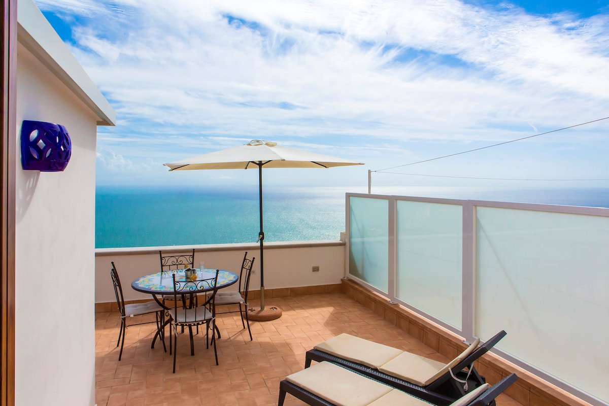 A spacious terrace offers an uninterrupted view of the sea, featuring a round dining table with chairs, a large umbrella, and two lounge chairs. The combination of sunlight and the ocean creates a serene outdoor environment.