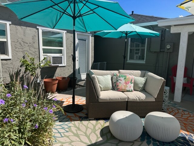 An inviting outdoor living area is depicted, featuring a comfortable sofa with decorative cushions. Two large umbrellas provide shade, and colorful poufs are arranged nearby. Potted plants and vibrant flowers add a touch of greenery, and a patterned outdoor rug enhances the setting.