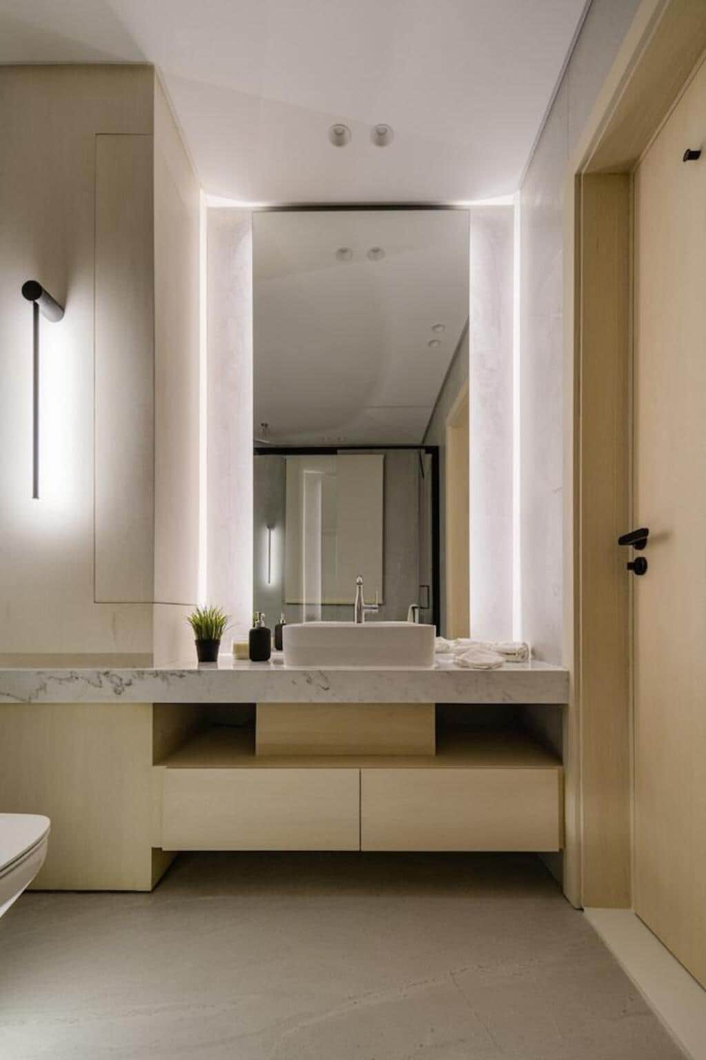 A modern bathroom features a sleek countertop accented with a small plant. Soft lighting highlights the contemporary mirror and minimalistic design elements, while a stylish faucet is positioned above the integrated sink. The overall color palette includes light tones, contributing to a clean and spacious appearance.