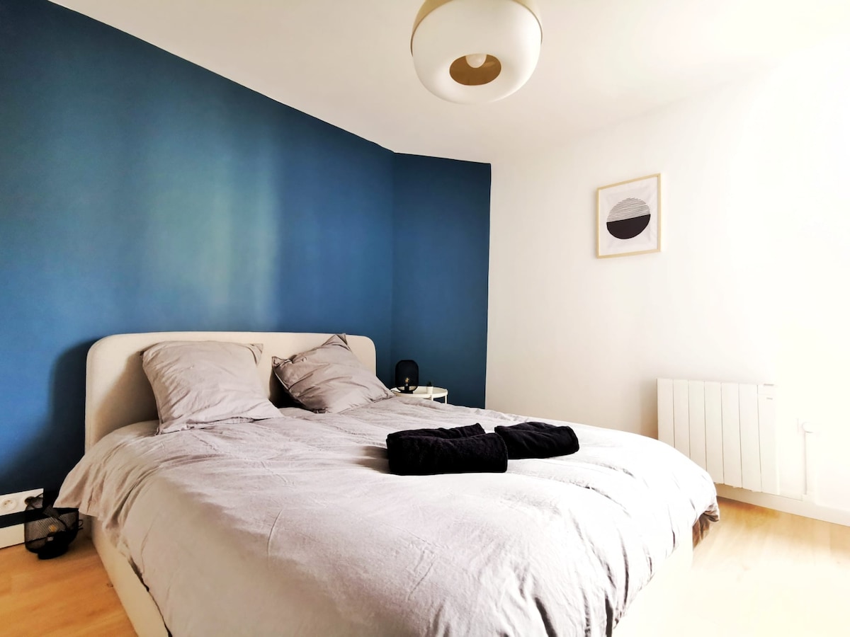 The bedroom features a spacious bed with neutral bedding and two black towels neatly placed on top. A blue accent wall provides a calming backdrop, complemented by a framed abstract artwork. A bedside table and radiator are also present, enhancing functionality.