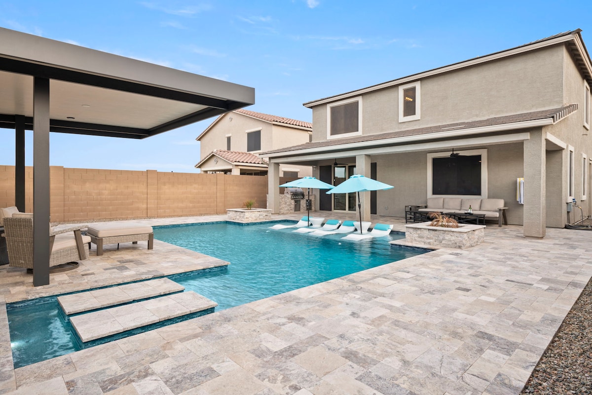 A modern backyard oasis is shown, featuring a heated saltwater pool surrounded by stone tiles. Comfortable outdoor seating under a covered patio offers shade, while two umbrellas provide additional sun protection over lounge chairs positioned by the pool. A fire pit area can be seen in the corner.