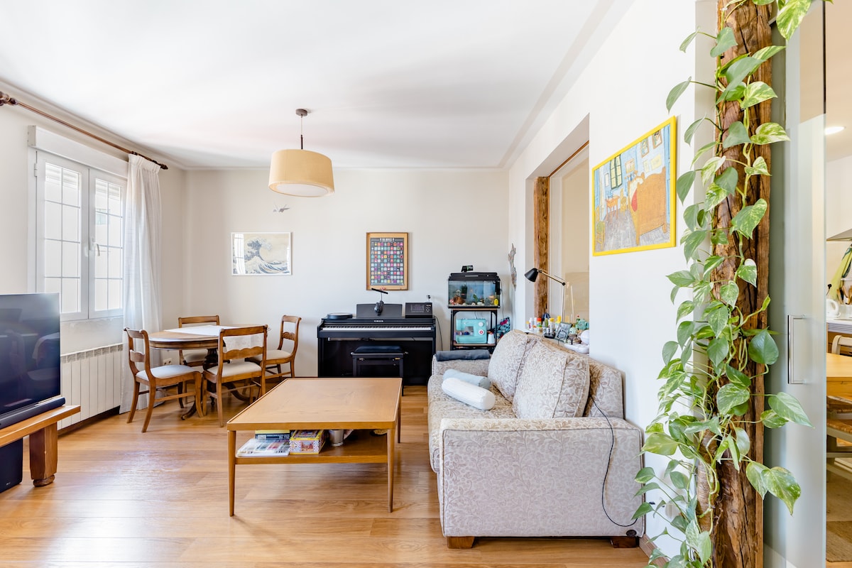 A spacious living area is illuminated by natural light through large windows. A wooden dining table with chairs is positioned next to a sofa and a coffee table. A piano and an aquarium are visible, contributing to a cozy and functional setting.
