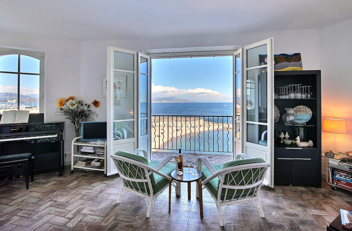 Two comfortable chairs are positioned near an open set of French doors, providing unobstructed views of the sea and beach. A small table is placed between the chairs, inviting relaxation. Soft natural light illuminates the room, highlighting the inviting atmosphere.