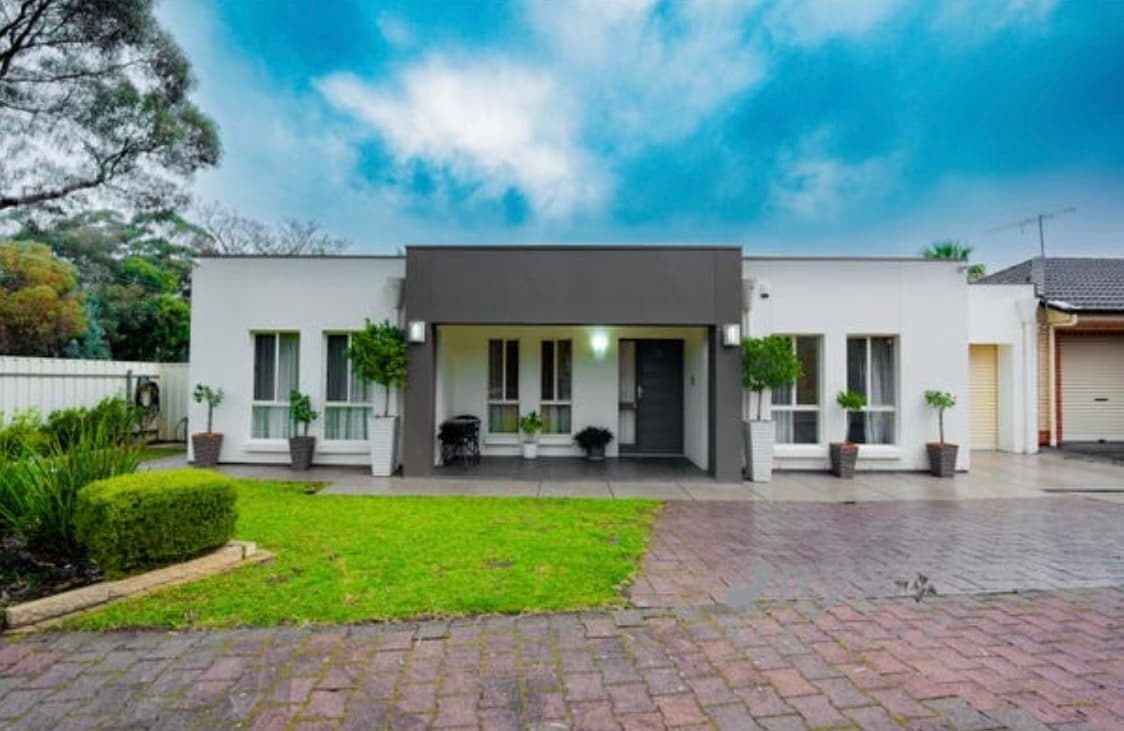 The front facade of the home is presented with a contemporary design, featuring large windows framed by neatly trimmed plants. A small porch is visually accentuated by a modern door. The driveway is paved, leading to a lock-up garage, with a backdrop of lush greenery.
