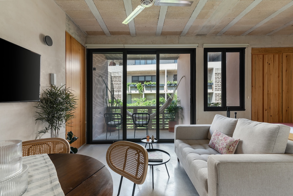 A spacious living area features large glass doors leading to a balcony, allowing natural light to enter. A comfortable sofa and a round dining table are visible, along with wicker chairs that complement the minimalistic decor. Greenery is observed outside through the glass doors.