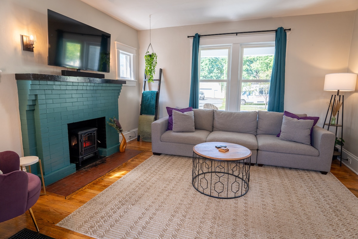A cozy living room is displayed, featuring a light gray couch positioned across from a retro-style fireplace. A round coffee table with a geometric design sits at the center. Natural light filters through two large windows, complemented by teal curtains, enhancing the inviting atmosphere.