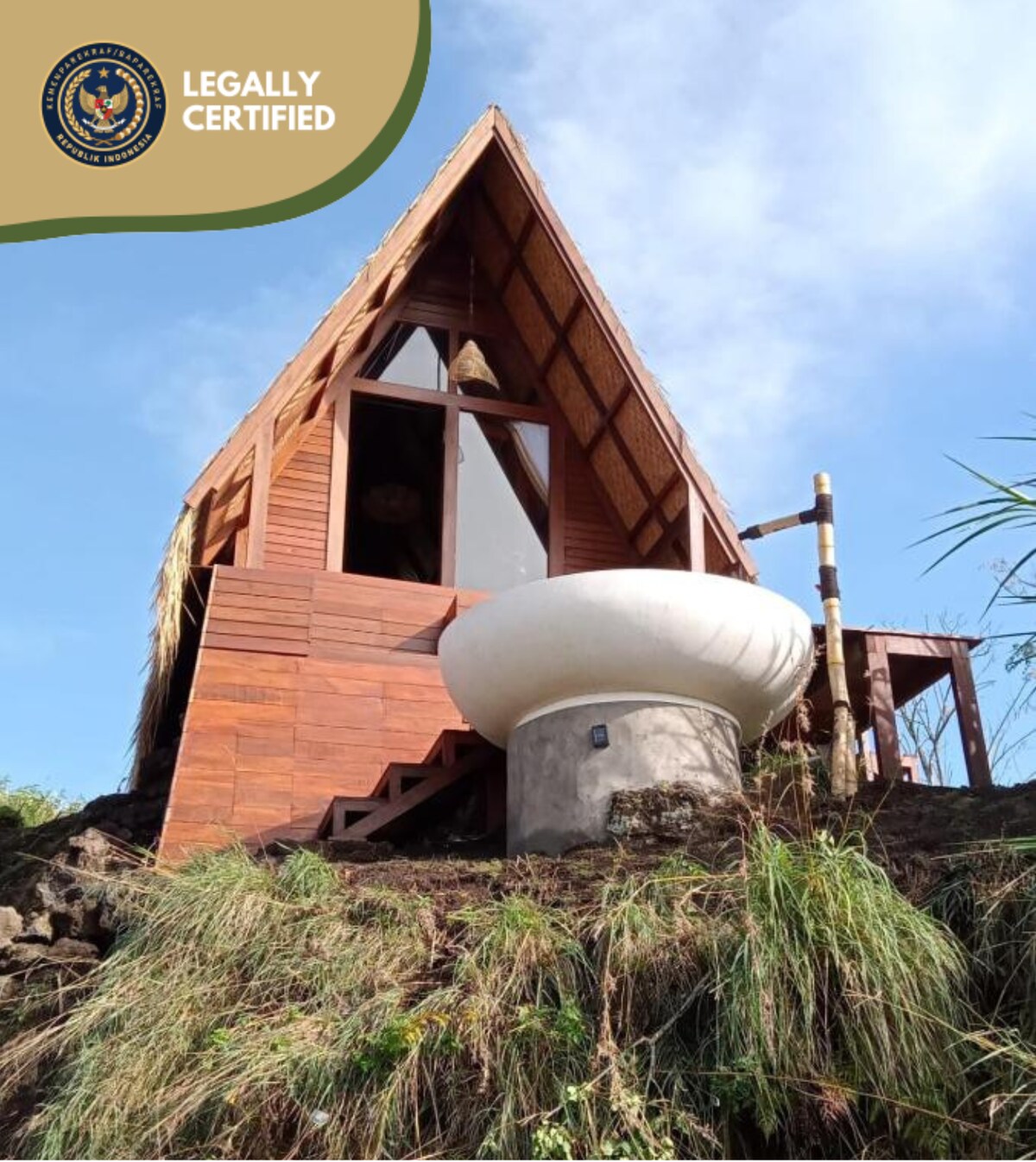A wooden bungalow features a sloped roof and large windows allowing for natural light. A round jacuzzi tub is located at the base, set against a backdrop of lush greenery. Steps lead up to the entrance, enhancing the rustic charm of the eco-friendly design.