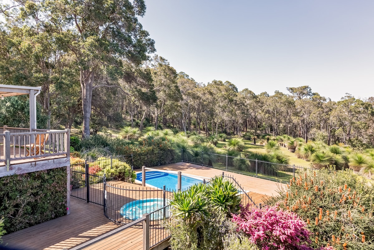 A serene outdoor space is displayed, featuring a sparkling pool surrounded by lush greenery and colorful flowering plants. The wooden deck leads to the pool area, which offers an expansive view of the tranquil bushland beyond.