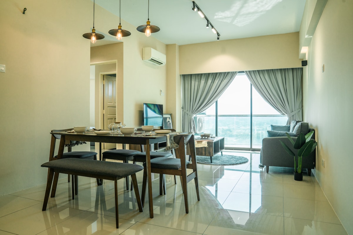 A modern dining area features a sleek wooden table surrounded by upholstered benches. Natural light floods the space through large windows, with a comfortable seating area visible in the background. Soft curtains frame the windows, enhancing the airy feel of the room.