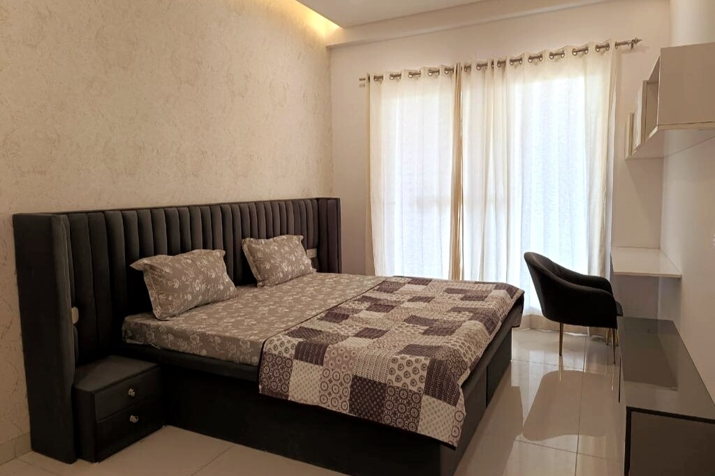 A modern bedroom features a spacious bed with a patterned blanket, complemented by two plush pillows. Natural light filters through sheer curtains at the window. A sleek chair sits beside a minimalist desk, enhancing the room's functionality.