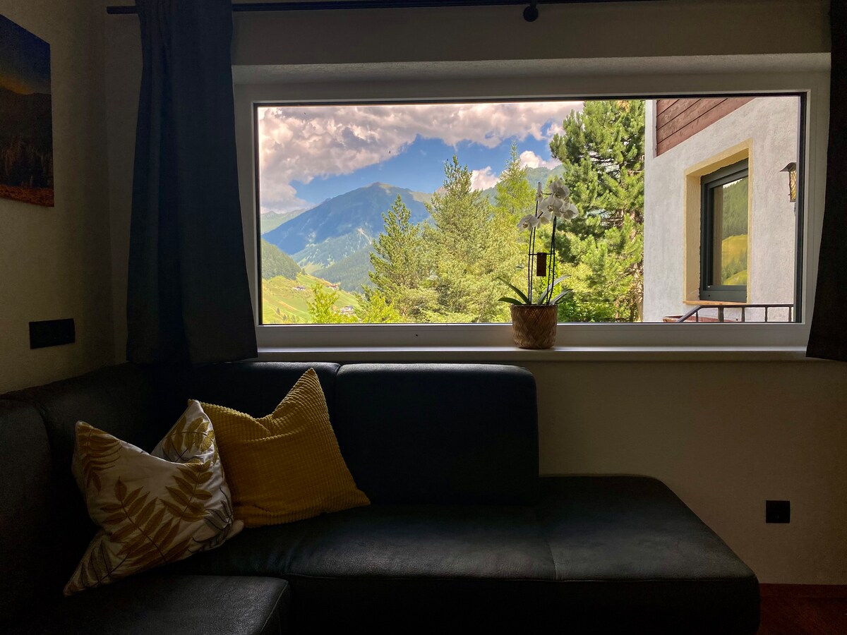 A cozy seating area is presented next to a large window, framing expansive mountain views. A small plant sits on the sill, while decorative pillows add a touch of comfort to the dark sofa. Natural light illuminates the space, enhancing the serene atmosphere.