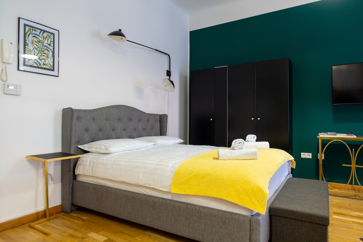 A spacious bedroom features a king-size bed with a light-colored duvet and a vibrant yellow throw. A small side table and a bench are visible beside the bed. The room is complemented by dark wardrobes and a mounted TV on the wall, with a pop of green on one wall.
