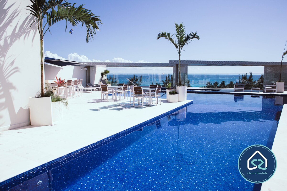 A spacious rooftop terrace features a heated pool with a vibrant blue tile surface. Lounge chairs are positioned along the edge, offering ocean views. Lush palm trees add greenery to the area, complemented by a seating arrangement, creating an inviting outdoor space.