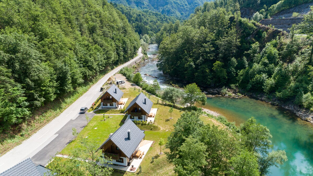 A serene landscape is visible, featuring three holiday houses set amidst lush greenery near the Kolpa River. A winding road runs alongside the river, which reflects the surrounding natural scenery. Gentle slopes rise in the background, enhancing the tranquil atmosphere.