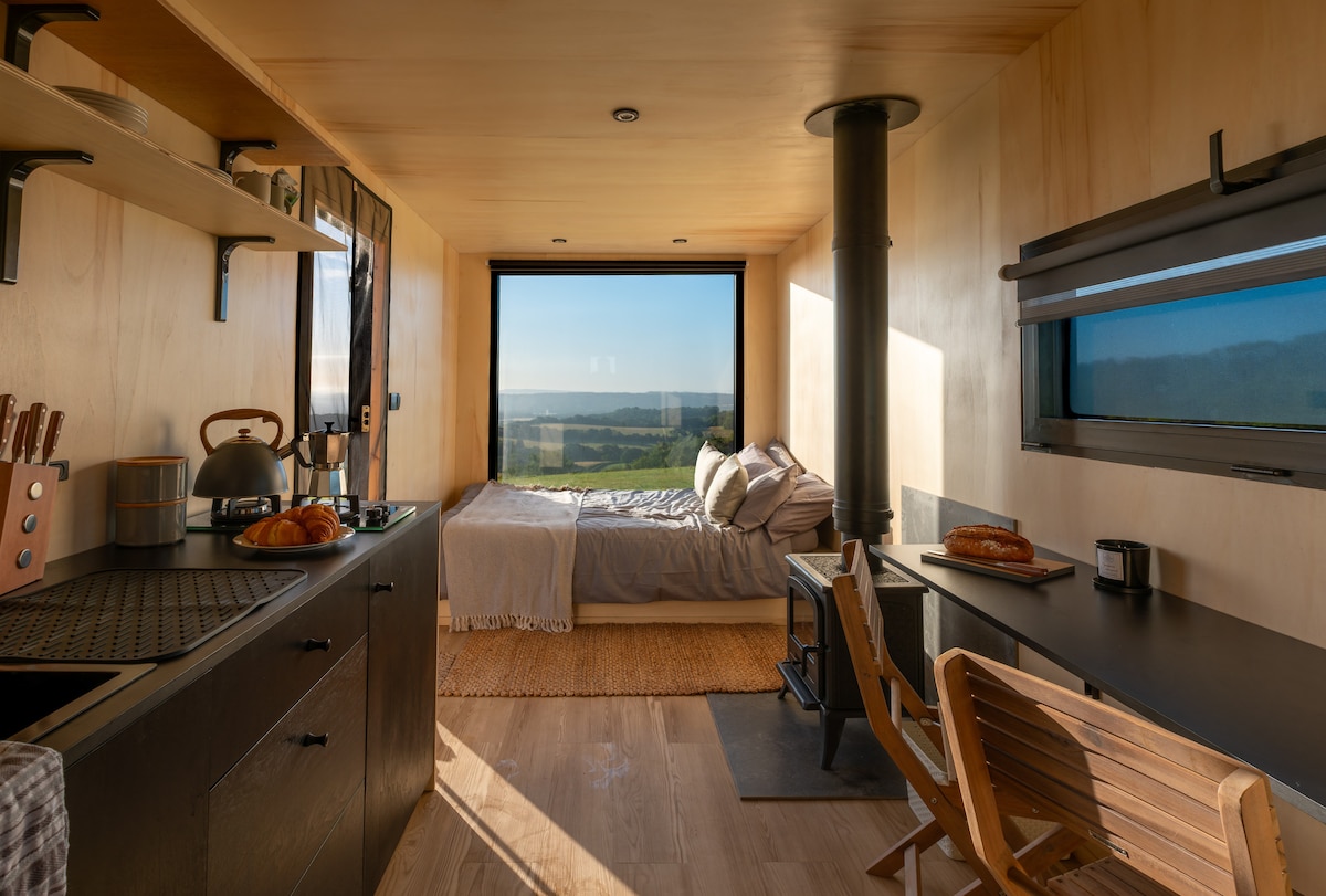 An interior view showcases a studio-style layout featuring a spacious bed near a large window, providing expansive views of the landscape. A well-equipped kitchen area includes a coffee maker and stove, while a small dining table and two chairs complement the cozy space.