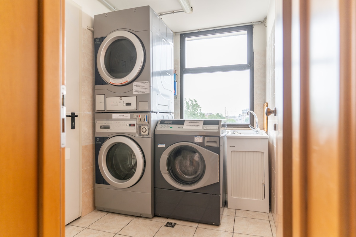 A laundry area features several modern appliances, including a stacked washer and dryer, along with a separate washing sink. Large windows provide natural light, creating a bright environment. The tiled floor enhances the functionality of the space.
