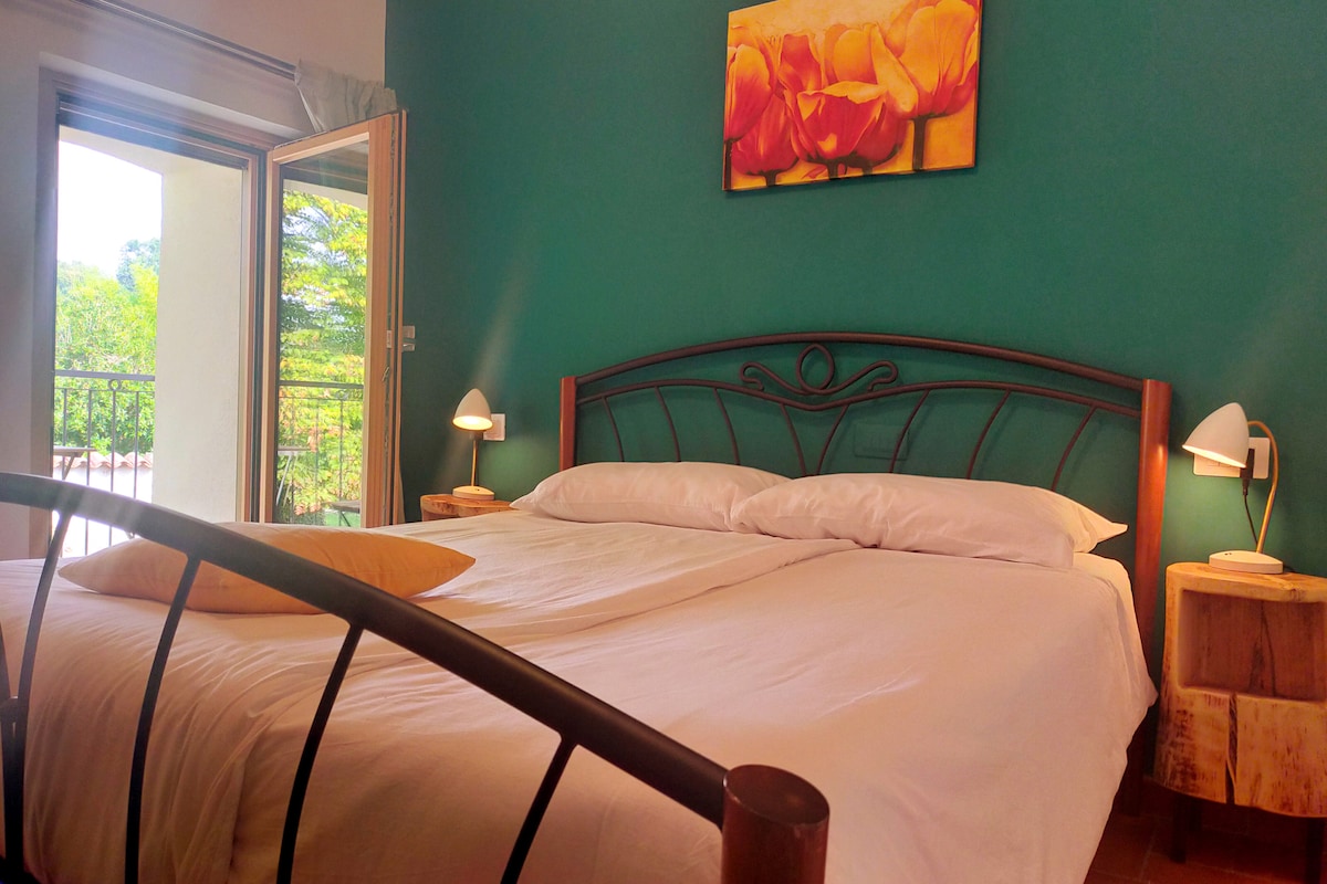 A spacious sleeping area features a large bed with crisp white linens. Two bedside lamps provide gentle lighting, complementing the green wall adorned with a floral artwork. Accessible doors lead to a balcony that overlooks the surrounding nature.