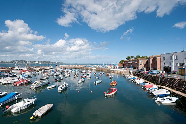 A vibrant marina scene features numerous boats anchored in calm waters. The shoreline is lined with colorful buildings, showcasing a blend of residential and commercial spaces. A clear blue sky with scattered clouds enhances the coastal atmosphere, inviting exploration of the nearby waterfront.