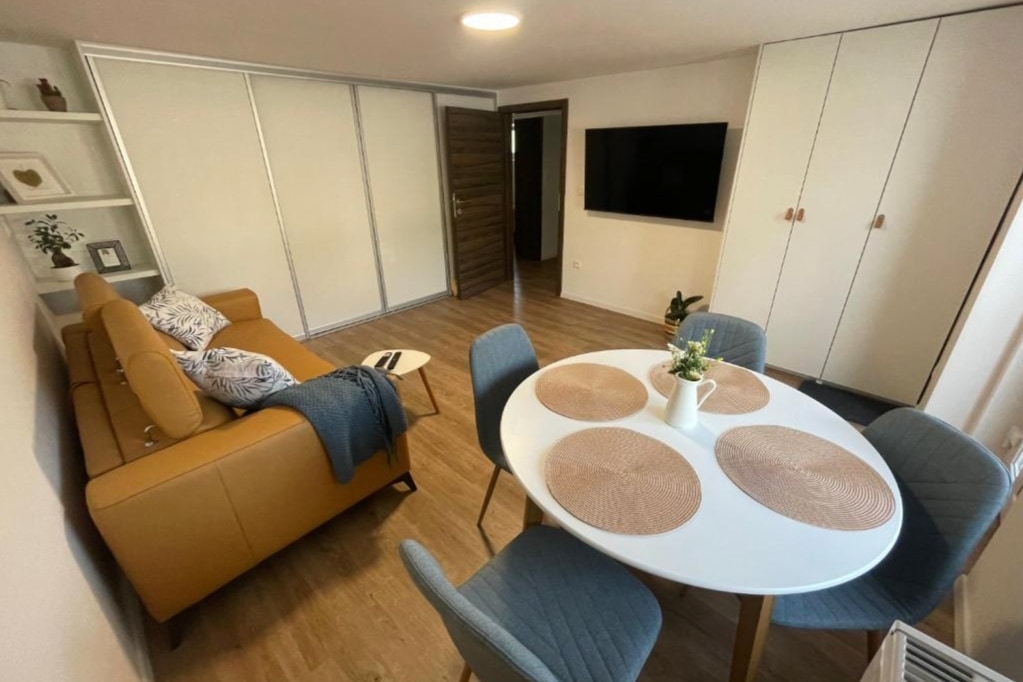 A bright living space features a leather sofa, complemented by decorative cushions. A round dining table with blue chairs offers a cozy dining area. Light wood flooring enhances the warmth of the room, while a large TV mounted on the wall provides entertainment options.