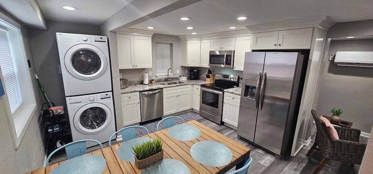 A modern kitchen with white cabinets and stainless steel appliances is visible. A dining table with light blue chairs sits in front of the kitchen area. A stackable washer and dryer are positioned beside the sink, with soft lighting enhancing the clean, organized space.
