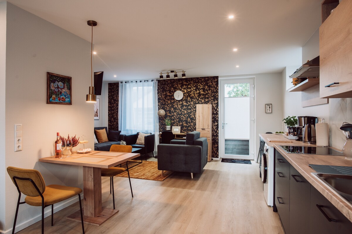 An inviting open-plan living area is displayed, featuring a dining table with two chairs near a cozy seating arrangement. Natural light filters through large windows, illuminating the modern kitchen equipped with essential appliances. A sleek door provides access to the outdoor space.