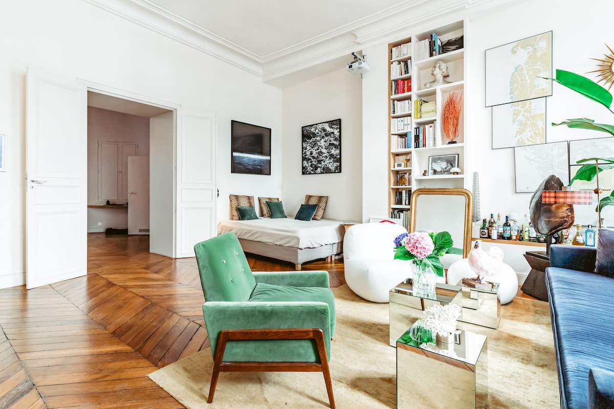 A spacious living area features a mix of seating options, including a turquoise upholstered chair and a large blue velvet sofa. A small glass coffee table with floral decor sits centrally, while bookshelves and artwork adorn the walls. A light wood floor enhances the openness of the space.