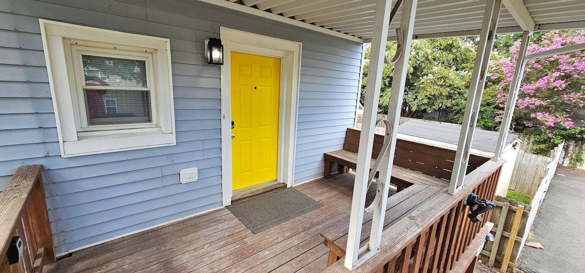 The entrance features a bright yellow door framed by a light blue exterior. A wooden porch with bench seating extends along the front, and blooming trees can be seen in the background, adding natural beauty to the welcoming entrance.