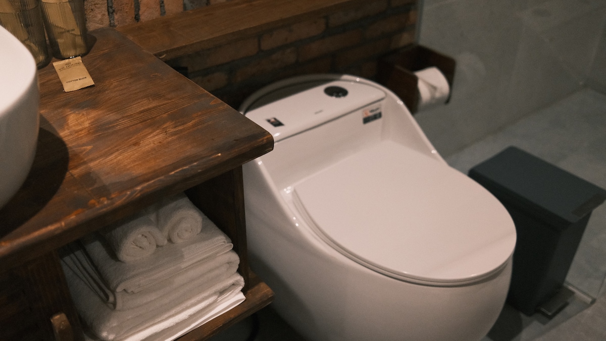 A modern bathroom features a sleek, high-efficiency toilet alongside a wooden counter with neatly stacked, white towels. A dark, discreet trash bin is positioned nearby, enhancing the functional design of the space.