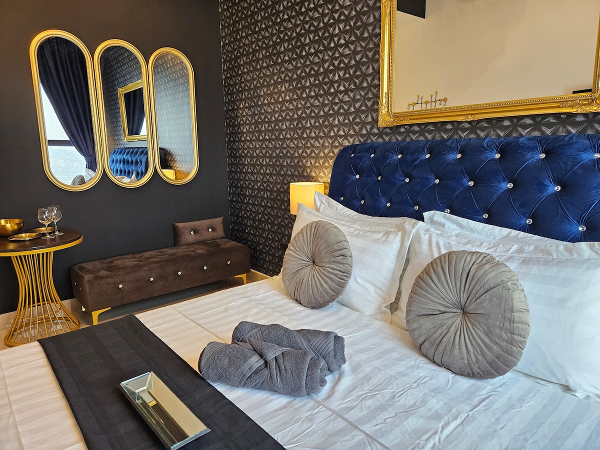 The bedroom features a queen bed dressed in soft linens with four plush pillows. A stylish blue tufted headboard and three mirrored accents are showcased on the dark geometric wallpaper. A cozy seating area with a small couch and a side table is also visible, offering a touch of elegance.