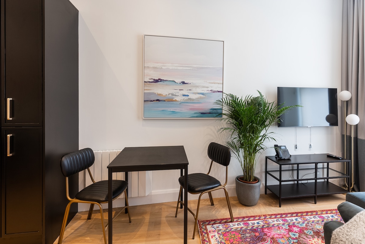 A minimalist living area is highlighted by a black table and two striped chairs. A colorful rug adds warmth to the space, while a large abstract painting adorns the wall. A potted plant brings a touch of greenery, complementing the sleek TV stand.