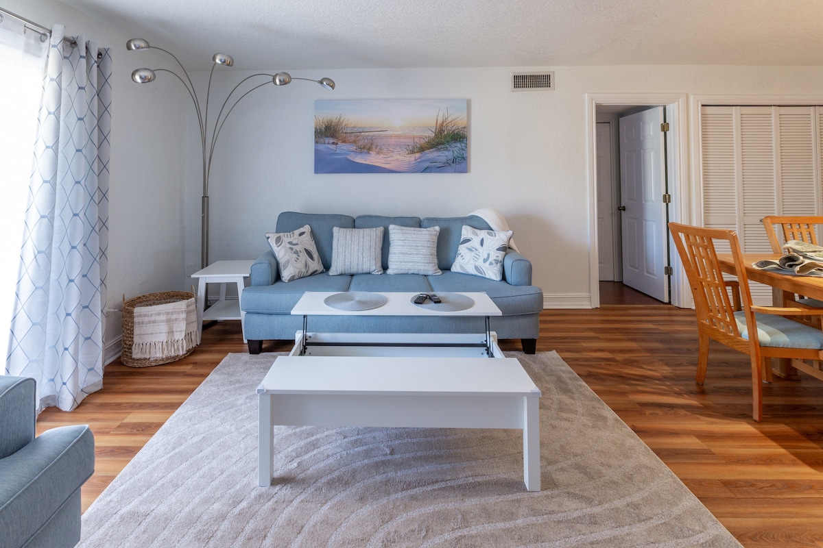 The living area is furnished with a light blue sofa adorned with decorative pillows. A white coffee table sits in front of the sofa, and a soft rug covers the floor. Natural light flows through a window draped with sheer curtains, enhancing the inviting environment.