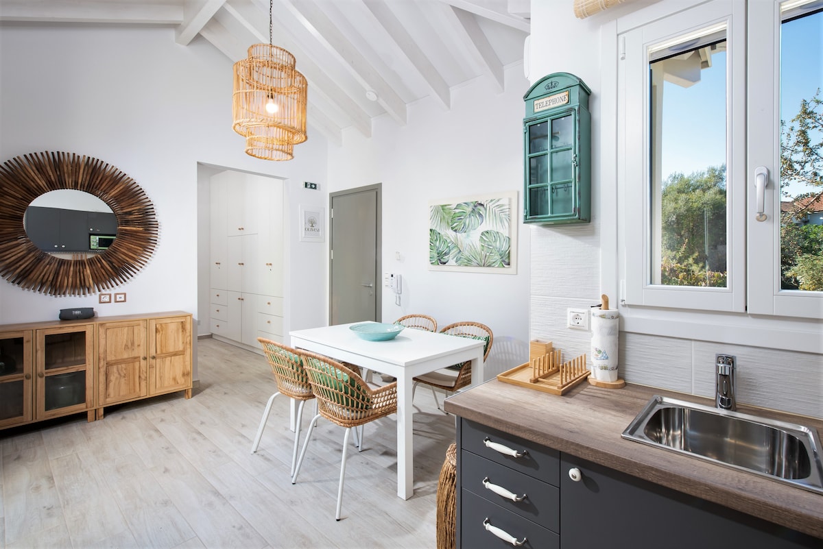 An open-plan dining area features a white table surrounded by four rattan chairs. A large round mirror adorns the wall, while light filters through a window, illuminating elements like a wooden storage cabinet and a green vintage wall cabinet.
