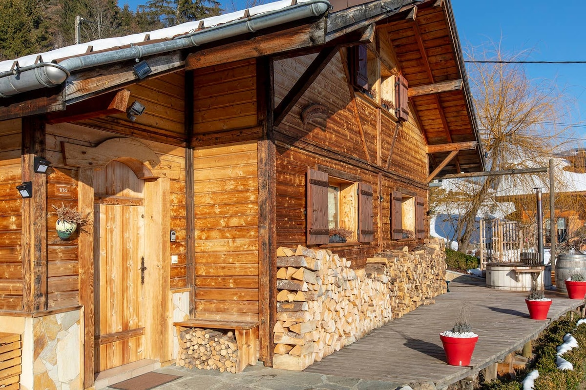 The exterior of the chalet features rich wooden textures and a welcoming front door. Stacked firewood is neatly arranged next to the entrance, and red planters add a pop of color to the surroundings. Bright sunlight enhances the natural hues of the building.