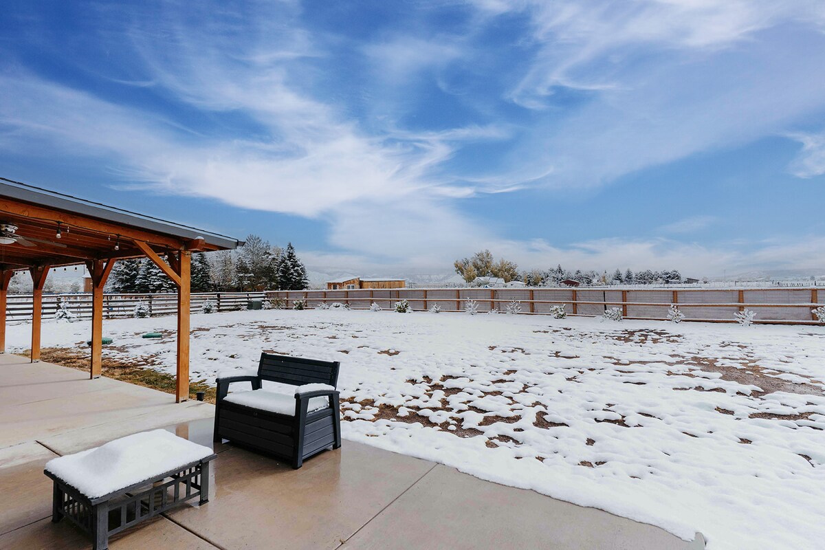 An outdoor space is visible, featuring a covered patio with wooden beams. A black seating area is positioned next to a fire pit, both sitting on a concrete surface. A snow-covered yard extends in the background, framed by a serene winter sky.