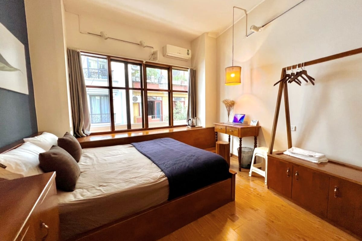 A private room features a spacious bed adorned with neutral bedding. Natural light floods the area through large windows, offering a view of the outside. A wooden desk is complemented by a chair and a rack for hanging clothes. The warm wooden tones create a cozy ambiance.