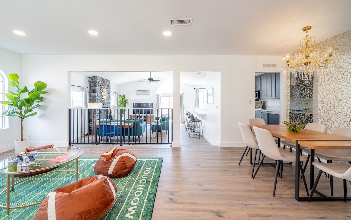 An open living space showcases a dining area with a wooden table and upholstered chairs, alongside a lounge area with bean bags positioned on a themed rug. A decorative chandelier hangs from the ceiling, while large windows allow natural light to brighten the room.