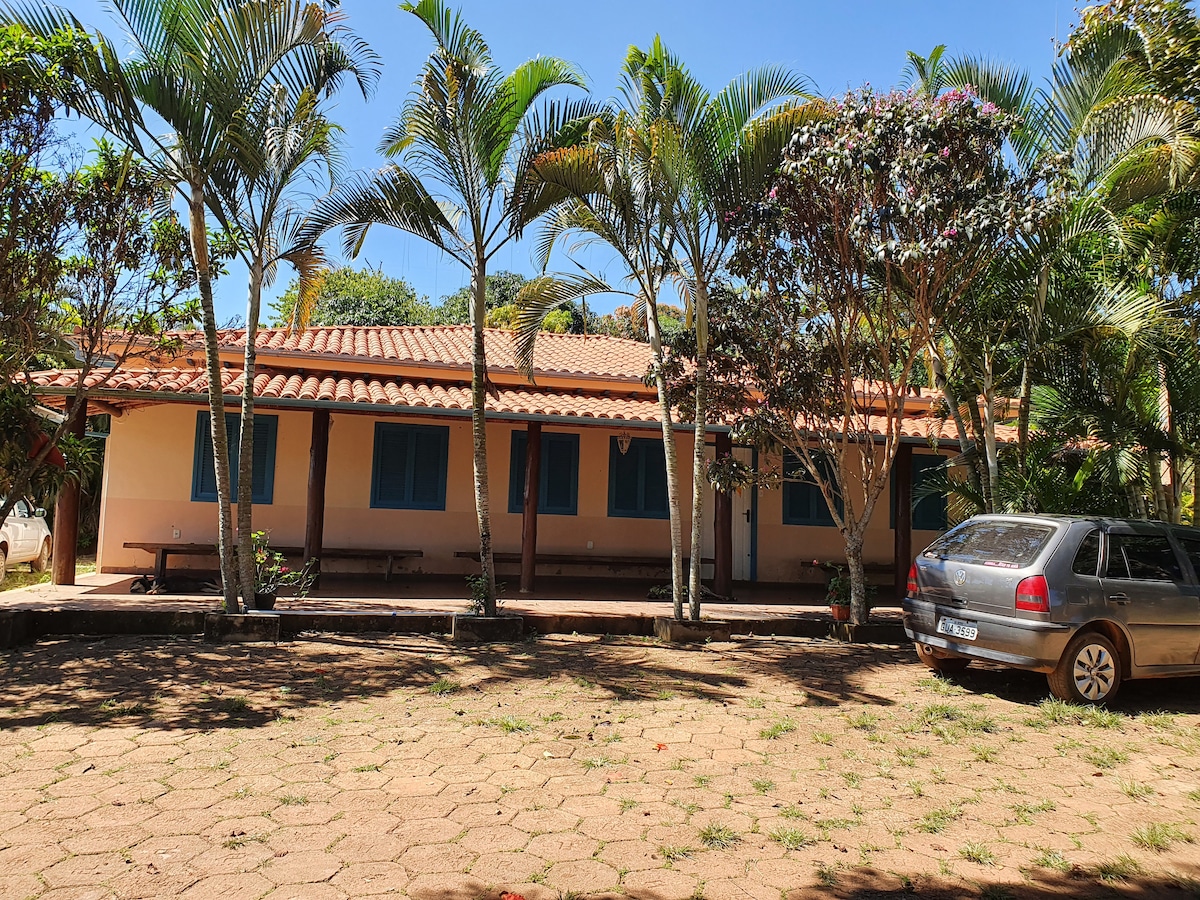 A single-story building is framed by several palm trees and flowering shrubs. The structure features a tiled roof and blue shuttered windows. A parked car is visible in the foreground on a stone-paved area, creating a welcoming approach to the entrance.