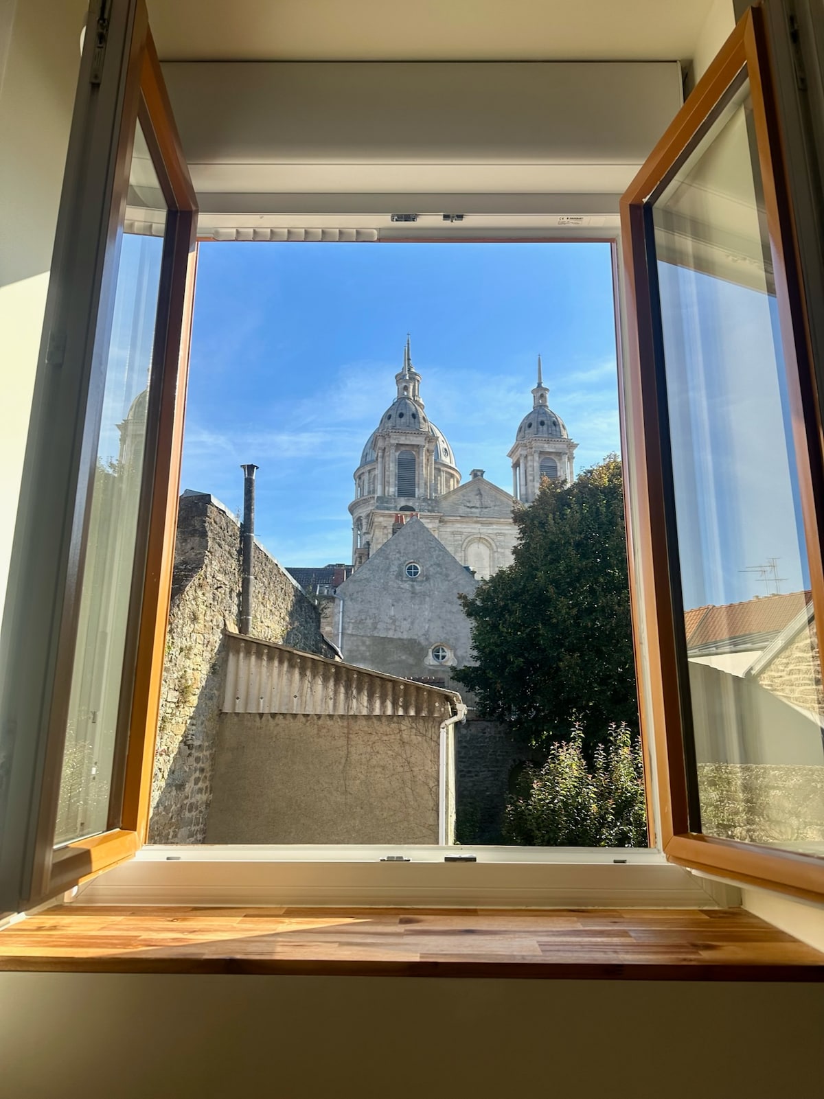 A view from an open window reveals a historic building with twin spires set against a clear blue sky. Surrounding architecture includes stone walls and greenery, inviting a sense of the local environment.