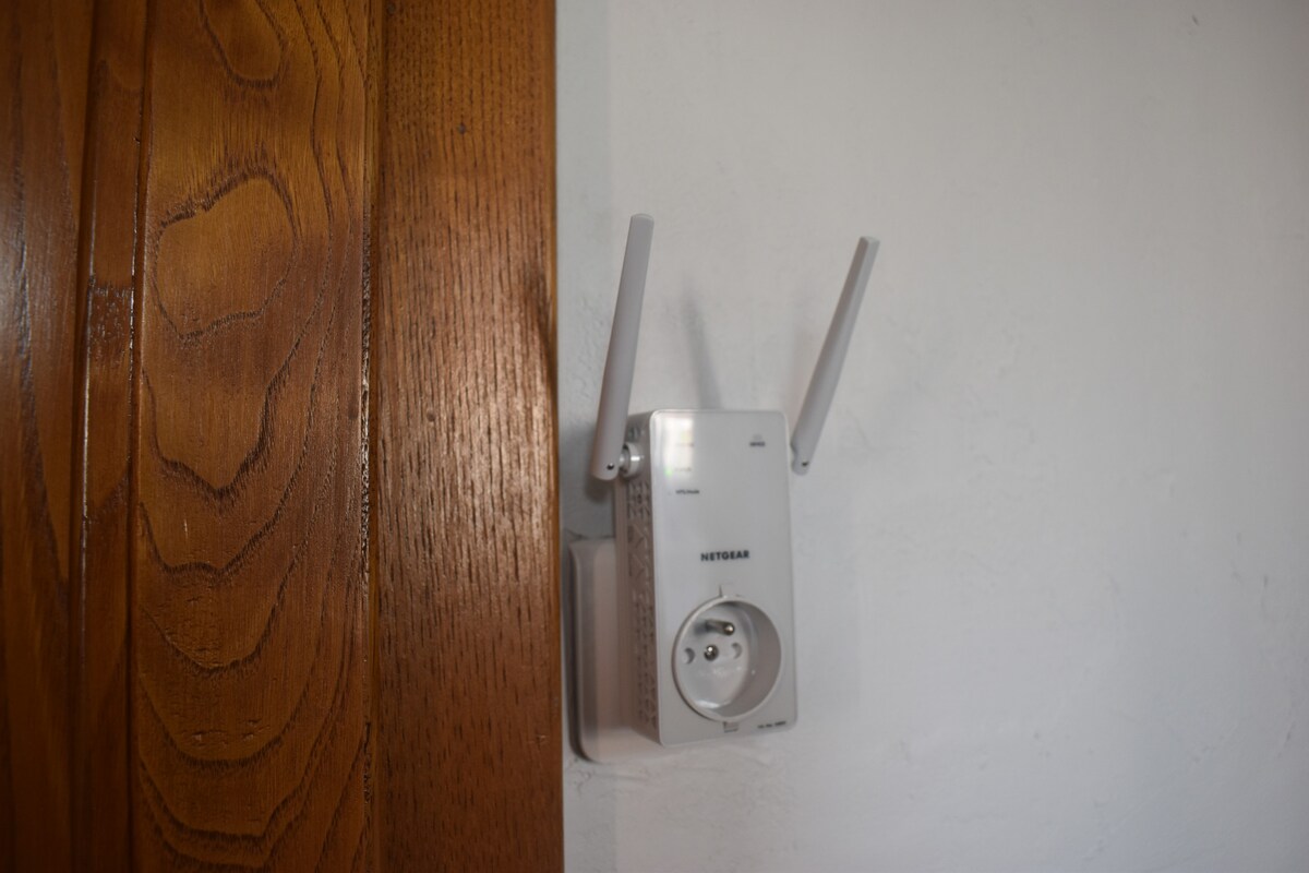 A wireless signal amplifier is mounted on a wall beside a wooden door. The device features two antennas extending outward and a plug socket integrated at the bottom, ensuring enhanced connectivity for remote work.