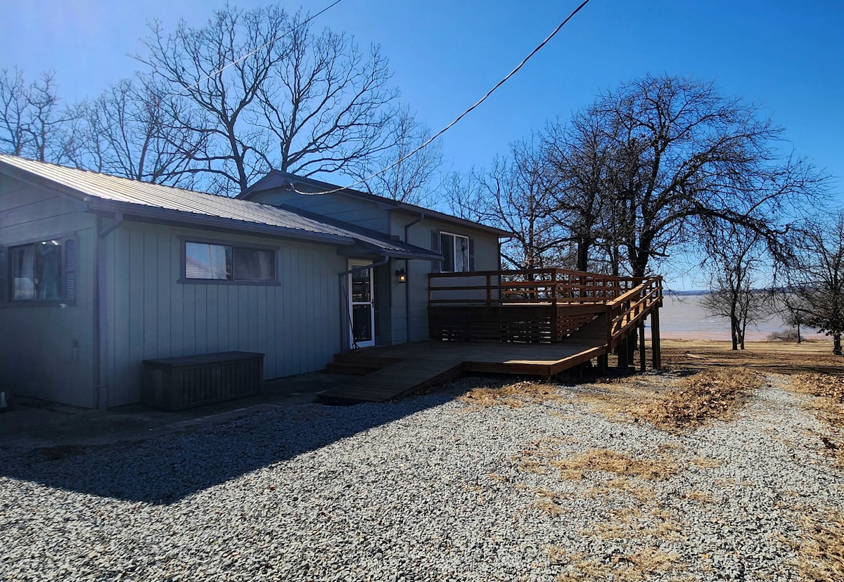 The exterior of a tri-story lakehouse is shown, featuring a wooden deck with a ramp for accessibility. The gravel driveway leads up to the entrance, framed by trees and open fields, offering views of the surrounding natural landscape.