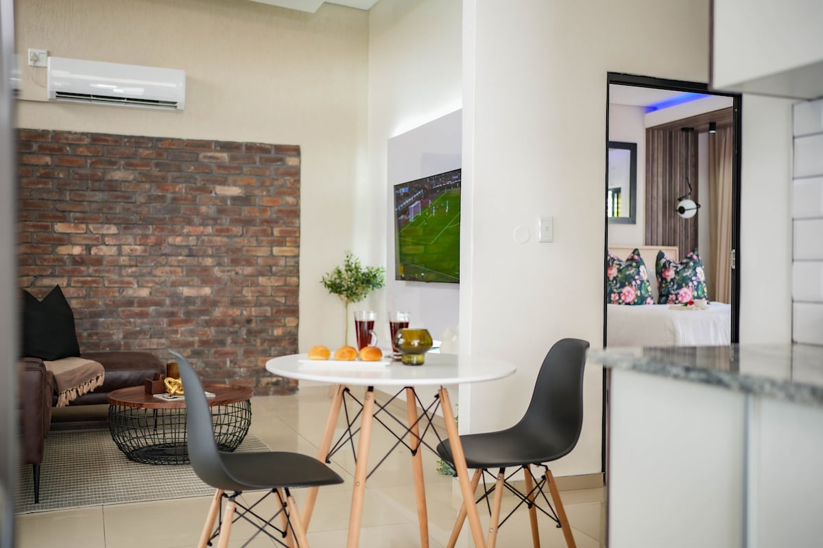 A modern dining area features a round table with two black chairs, accompanied by a bowl of pastries. A textured brick wall serves as a backdrop, while a flat-screen TV is mounted nearby. The living space is open to a cozy seating area with a brown couch.