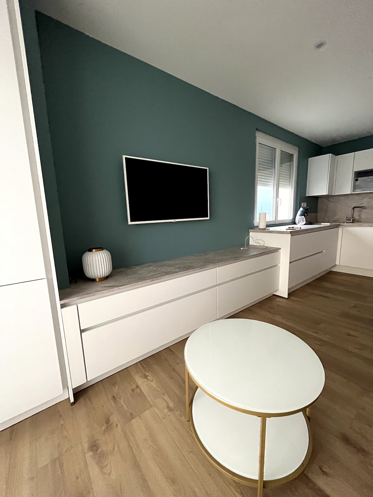 A modern living area features a light wood floor and a soothing green accent wall. A white entertainment unit houses a flat-screen TV, while a circular coffee table with a white top and gold base sits in the center. The adjacent kitchen area is equipped with contemporary cabinetry.